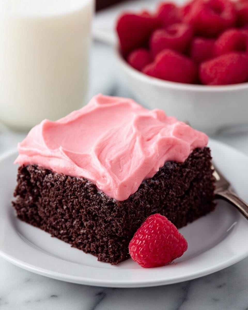 A square piece of chocolate cake with one smooth, thick layer of bright pink frosting on top sits on a white plate. The cake layer is dark brown, moist, and textured, while the pink frosting is creamy with soft swirl marks. Next to the cake on the plate is a fresh, red raspberry. In the background, there is a white bowl filled with raspberries and a glass of milk, all set on a white marbled surface. Photo taken with an iphone --ar 4:5 --v 7