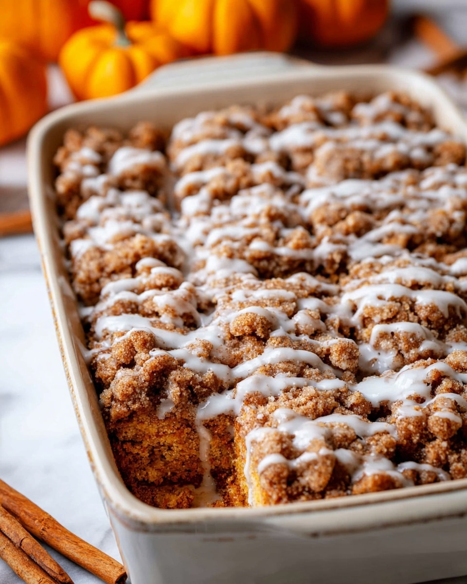 A close-up view of a multi-layered dessert in a white baking dish, featuring a crumbly top layer of golden-brown streusel with irregularly sized clumps covered in a light drizzle of white icing. The layers beneath are not visible but the texture on top shows rich, crunchy bits that contrast with the smooth, glossy glaze. The dish is set on a white marbled surface with cinnamon sticks and small pumpkins blurred in the background, adding a warm, cozy feel. The dessert is sliced into square portions, ready to be served. Photo taken with an iphone --ar 4:5 --v 7
