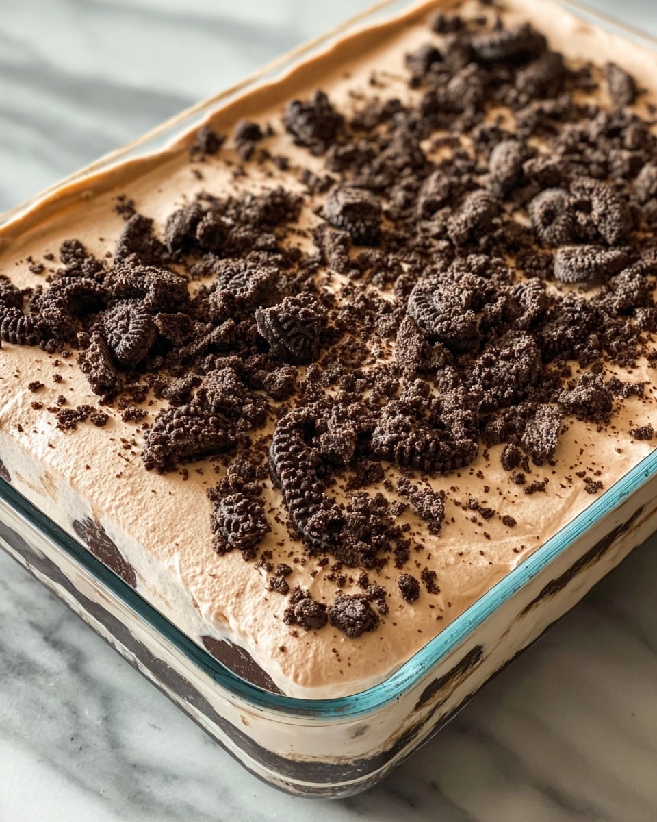 A close-up image shows a rectangular glass dish filled with a layered dessert. The bottom layer is not fully visible, but the top thick layer is light brown and smooth, resembling a creamy mousse or pudding. On top of this creamy layer, there is a generous amount of crushed dark chocolate cookies spread unevenly, with some crumbs and bigger chunks scattered around. The dish is set on a white marbled surface. photo taken with an iphone --ar 4:5 --v 7