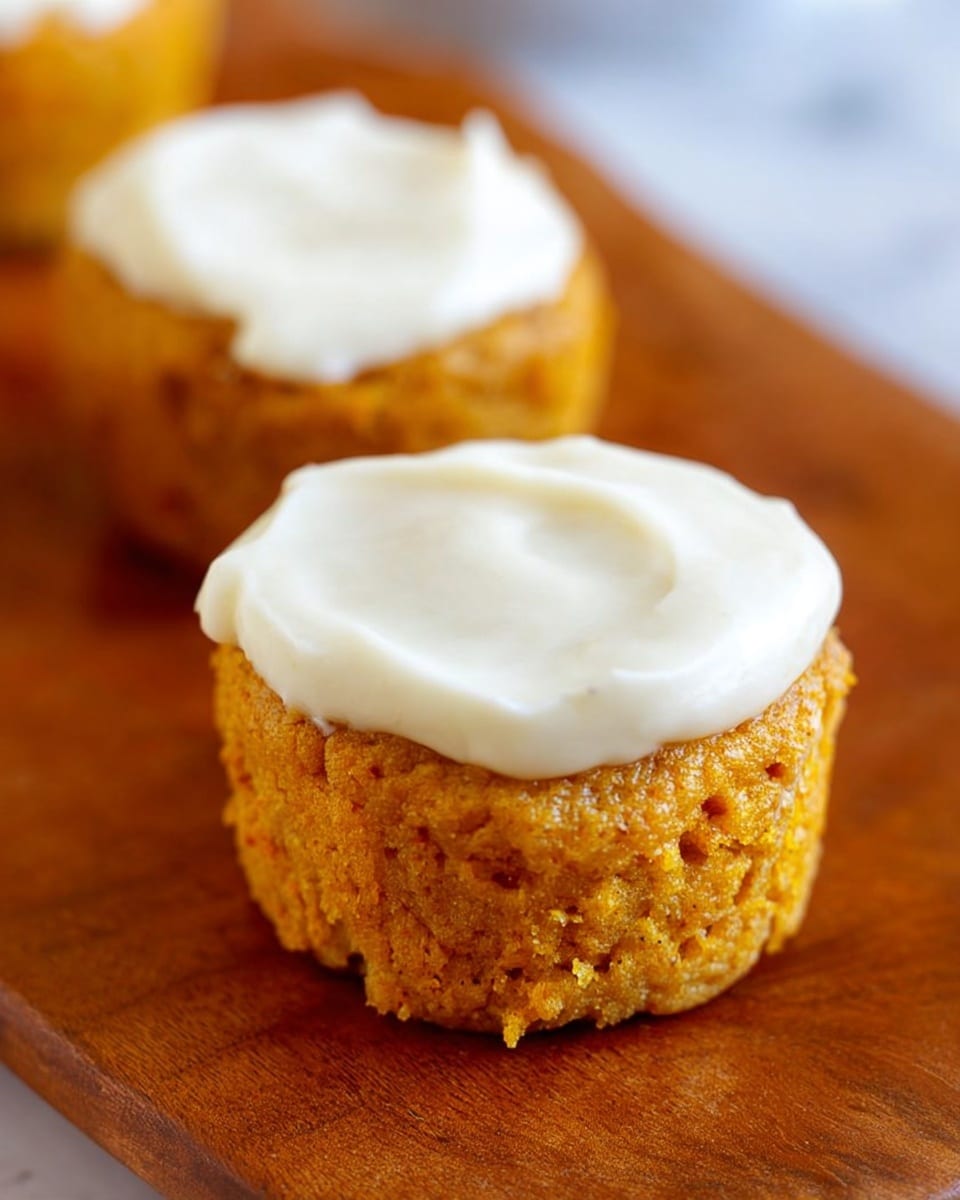 The image shows two small pumpkin cupcakes on a wooden surface, each topped with a smooth, thick layer of white cream cheese frosting that covers the top fully. The cupcakes have a moist and crumbly orange texture with tiny visible bits throughout, giving a homemade appearance. The frosting layer is evenly spread with soft swirls, adding a creamy contrast to the rough cupcake base. The cupcakes are close to each other, with one slightly behind the other, both centered in the image on a white marbled texture. photo taken with an iphone --ar 4:5 --v 7