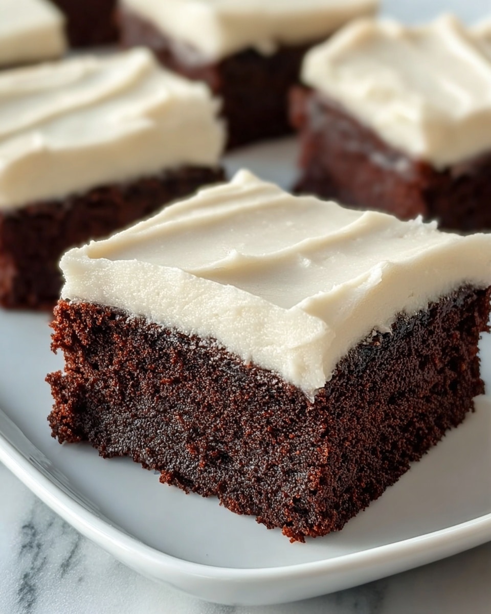 A close-up of square chocolate cake pieces with two visible layers: a thick, dark brown, moist and crumbly cake base topped with a smooth, creamy, off-white frosting layer that is evenly spread with gentle swirls. The cake pieces are placed on a white plate over a white marbled surface. The focus is on the front cake piece, with more pieces blurred in the background. photo taken with an iphone --ar 4:5 --v 7