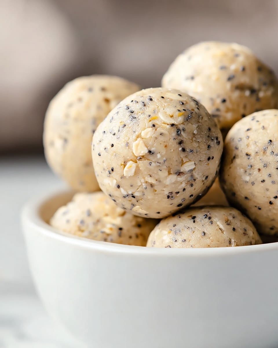 A close-up image of round energy bites stacked in a white bowl on a white marbled surface. The energy bites are light beige with visible small black poppy seeds and flakes of oatmeal scattered evenly on the surface, giving them a textured look. The focus is on one energy bite in the front, showing its smooth but slightly grainy texture with tiny dark seeds and oat pieces embedded throughout. The background is softly blurred, highlighting the detail on the energy bites in the front. photo taken with an iphone --ar 4:5 --v 7
