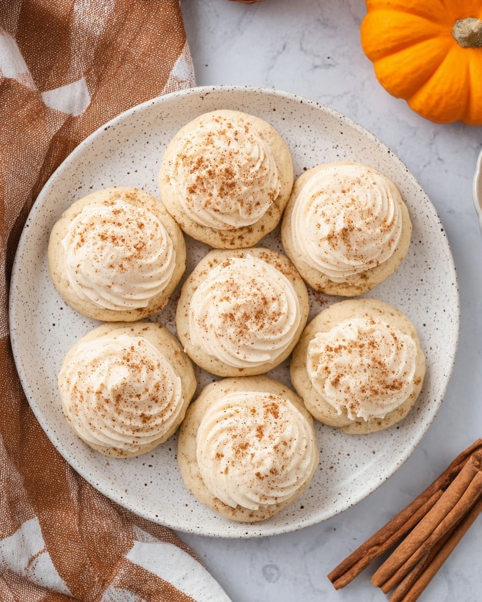 A white speckled plate holds eight round cookies with a light beige color, each topped with smooth swirls of creamy frosting. The frosting is sprinkled with a fine dusting of brown spice, giving a textured look. The cookies are arranged close together on the plate, which is set on a white marbled surface. To the side, there is a brown and white checkered cloth and two cinnamon sticks, adding a warm touch. In the background, a small orange pumpkin adds a pop of color. photo taken with an iphone --ar 4:5 --v 7