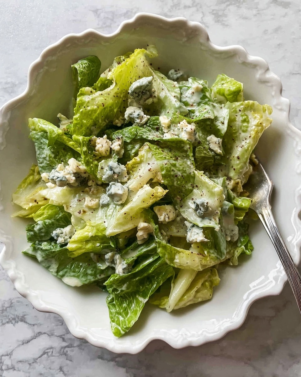 A close-up of a white scalloped ceramic bowl filled with a green leafy salad. The salad has fresh, crisp romaine lettuce leaves in various shades of light to medium green, torn and layered throughout. Scattered on top and mixed in are small, creamy white chunks of blue cheese with visible blue-green mold veins, adding texture and color contrast. There is a light, creamy dressing coating the lettuce and cheese, with some specks of black pepper and herbs sprinkled on top. A silver fork rests inside the bowl, partially visible on the right side. The bowl is placed on a surface with a white marbled texture. photo taken with an iphone --ar 4:5 --v 7