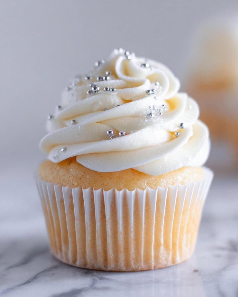 A close-up view of a single cupcake with a light golden brown base wrapped in a white pleated paper liner, topped with a swirl of smooth, creamy white frosting sprinkled with small shiny silver edible beads, all placed on a white marbled surface with a soft blurred background. Photo taken with an iphone --ar 4:5 --v 7