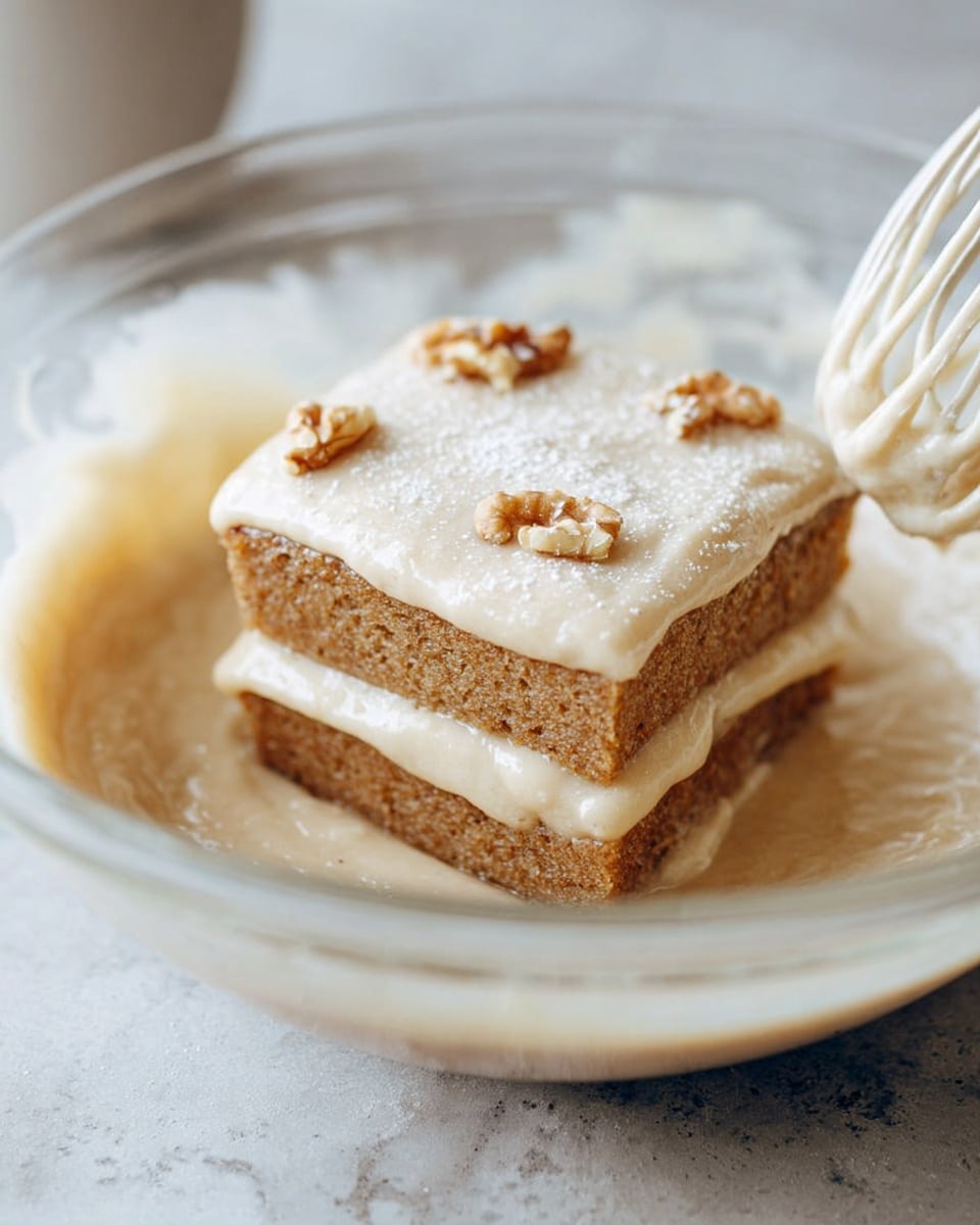 The image shows three stacked square pieces of banana cake on a white plate set on a white marbled surface. Each cake piece has two layers: a moist, light brown banana cake base with a slightly crumbly texture and a smooth, creamy white frosting layer on top. Thin banana slices are placed between the cake pieces and on the top layer. The top cake layer is garnished with walnut halves and a light dusting of powdered sugar, giving a delicate finish. The background is softly blurred with a cup in the distance, highlighting the stack of banana cake. Photo taken with an iphone --ar 4:5 --v 7