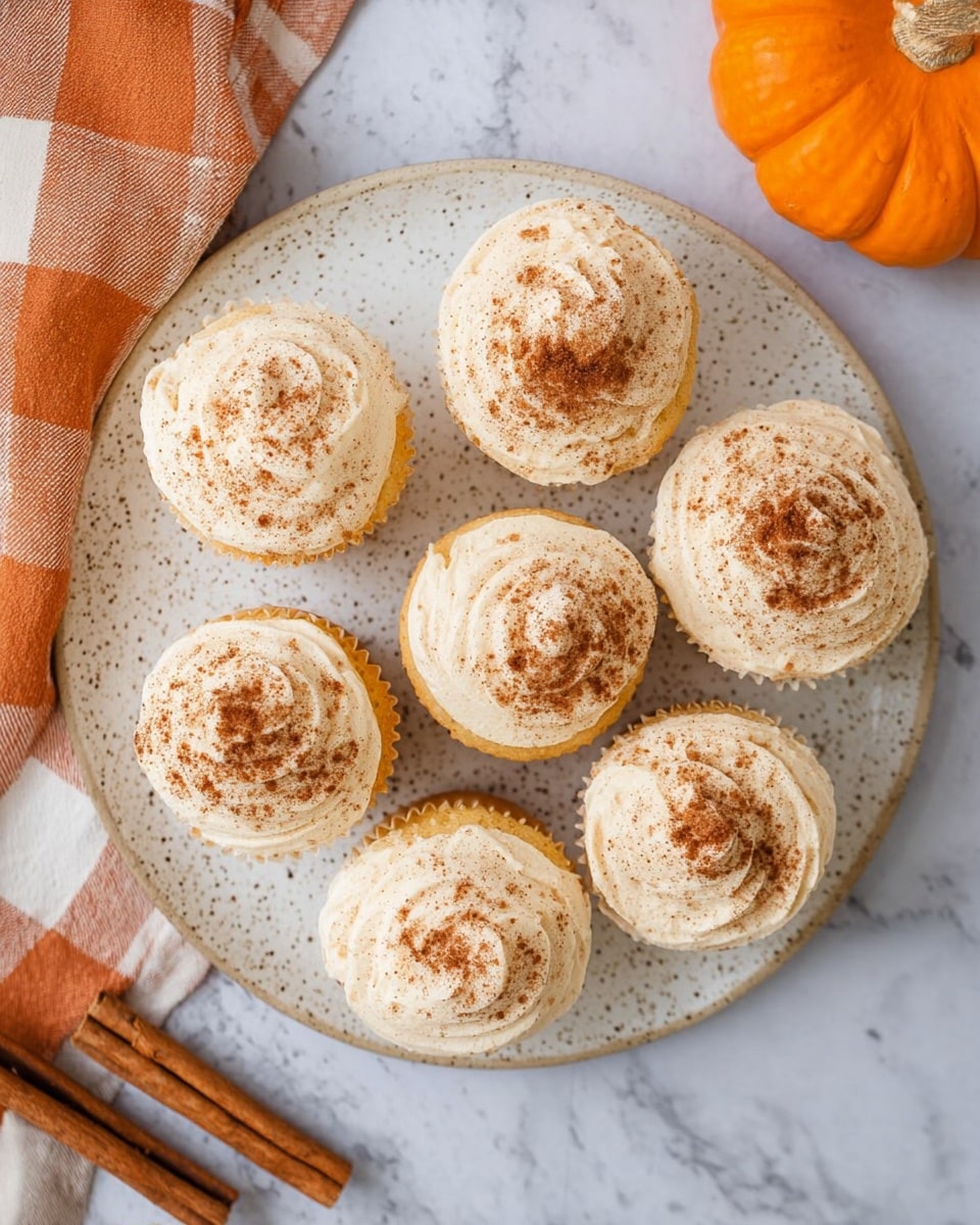Seven light beige frosted cupcakes are arranged evenly on a round white speckled plate, each cupcake topped with a dusting of brown cinnamon powder. The frosting is swirled on, giving a soft, creamy texture to each cupcake. The plate is placed on a white marbled surface, beside a folded orange and white checkered napkin on the left and two cinnamon sticks at the bottom left corner. In the upper right corner, a small orange pumpkin adds a touch of autumn color. photo taken with an iphone --ar 4:5 --v 7