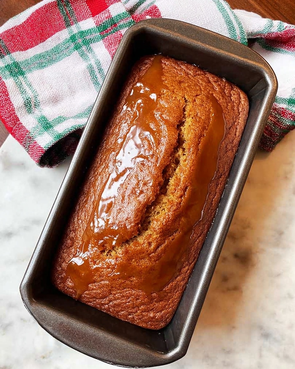 A freshly baked rectangular bread loaf sits in a dark metal baking pan, glossy with a thick caramel-colored glaze covering the top and pooling slightly around the edges. The bread’s surface is golden brown with a few cracks and a slightly rough texture, showing it is moist inside. The pan is placed on a white marbled texture surface with a plaid cloth in white, red, green, and blue stripes casually laid beside it. photo taken with an iphone --ar 4:5 --v 7
