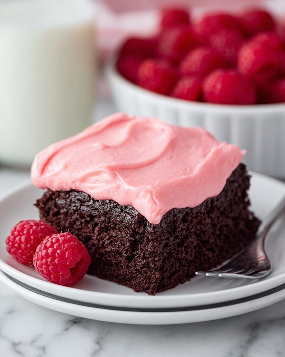 The image shows a close-up of a square chocolate cake piece with one thick layer of smooth, bright pink frosting covering the top. The cake itself is dark brown and looks soft and moist with a slightly crumbly texture on the sides. The cake sits on a white plate with a delicate pink flower pattern along the edge. Fresh red raspberries are placed beside the cake on the plate, adding a pop of color. A silver fork is placed on the left side of the plate. In the background, blurred, there is a white cup filled with raspberries and a clear glass of milk. The surface beneath everything has a white marbled texture. photo taken with an iphone --ar 4:5 --v 7