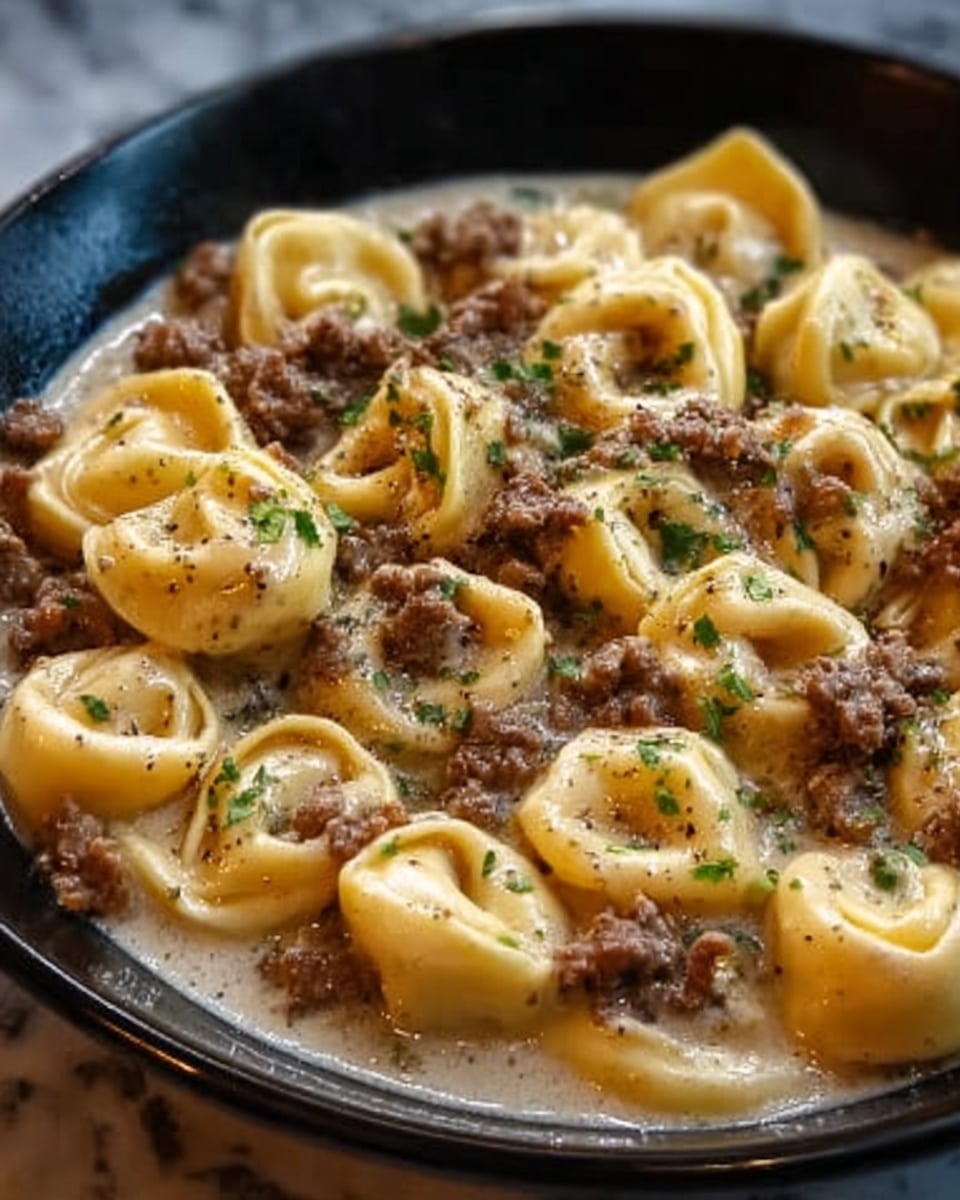 The image shows a close-up of a black bowl filled with pasta tortellini covered in a creamy sauce. The tortellini are folded into small round shapes with visible folds and a soft yellow color. On top, there are pieces of cooked ground meat that are dark brown, scattered evenly across the pasta. Small green herbs are sprinkled over the dish for garnish. The creamy sauce underneath the meat and pasta has a smooth texture and light color, blending softly with the pasta. The background is a white marbled surface. Photo taken with an iphone --ar 4:5 --v 7