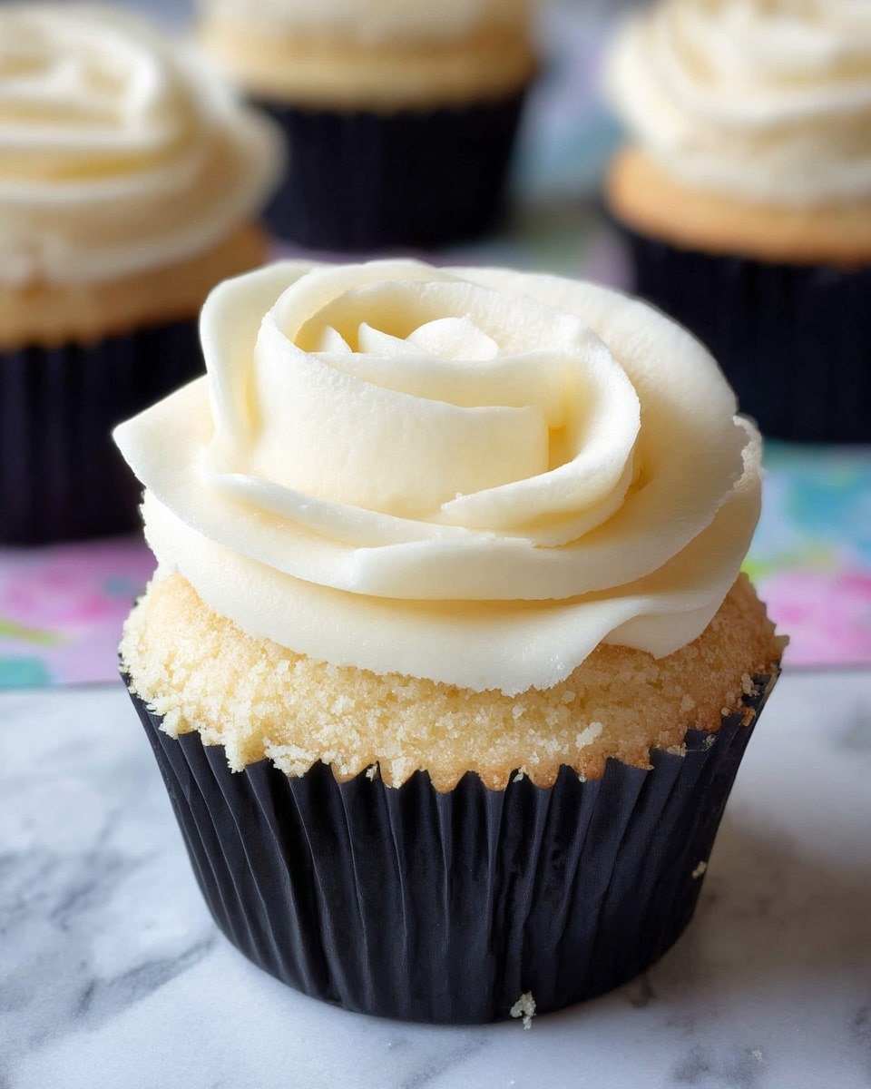 A close-up view of a single vanilla cupcake with one thick layer of white, creamy frosting piped in a swirl that looks like a rose on top. The cupcake cake layer is light yellow with a soft, spongy texture and small visible crumbs. The cupcake sits in a black paper liner on a white marbled surface with a soft focus view of other similar cupcakes in the background. Photo taken with an iphone --ar 4:5 --v 7