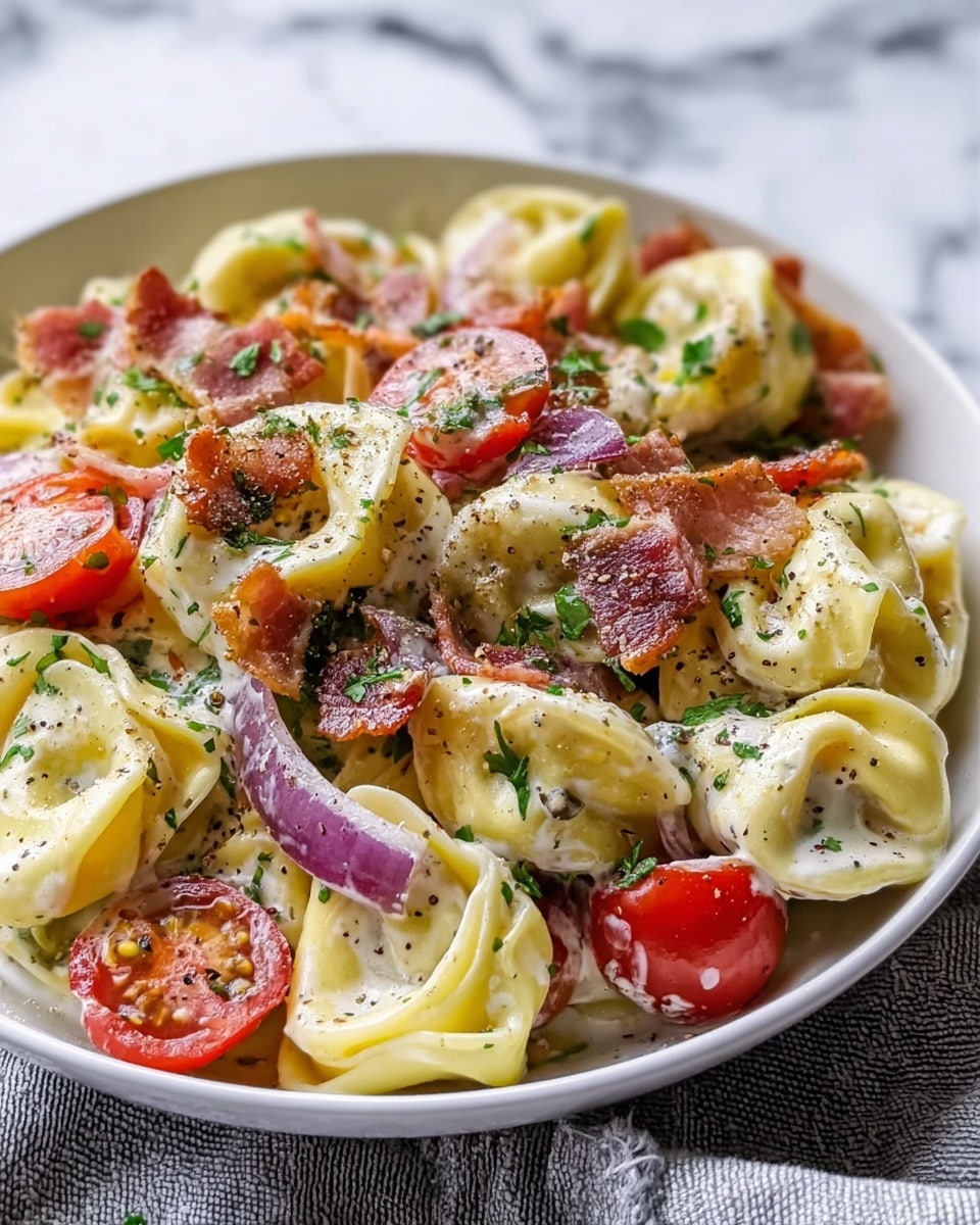 The image shows a white bowl filled with creamy tortellini pasta. The tortellini pieces are pale yellow and soft, coated evenly in a white creamy sauce. Scattered throughout the pasta are bright red cherry tomato halves, thin slices of purple red onion, and crispy brown bacon pieces. The dish is sprinkled with fresh green herbs and cracked black pepper, giving it a fresh and colorful look. The bowl sits on a white marbled surface with a light gray cloth underneath. Photo taken with an iphone --ar 4:5 --v 7
