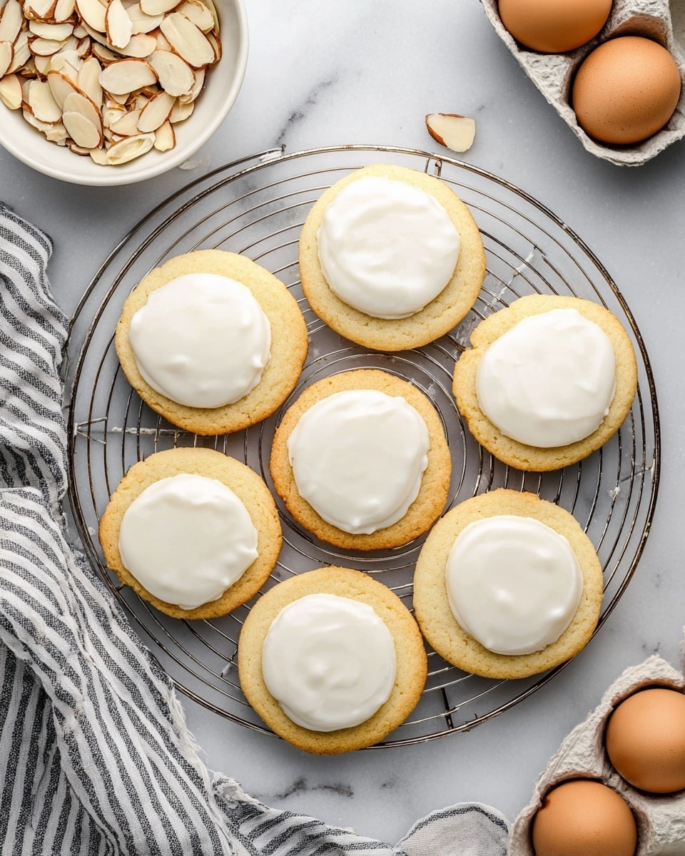 Seven round soft cookies with smooth white icing on top are arranged on a round metallic cooling rack placed on a white marbled surface. Each cookie has one layer of golden-brown dough with a clean, even coating of white icing centered on top. Nearby, a white bowl filled with sliced almonds and a white egg carton holding two brown eggs are visible. A striped cloth is partly seen in the lower left corner. Photo taken with an iphone --ar 4:5 --v 7