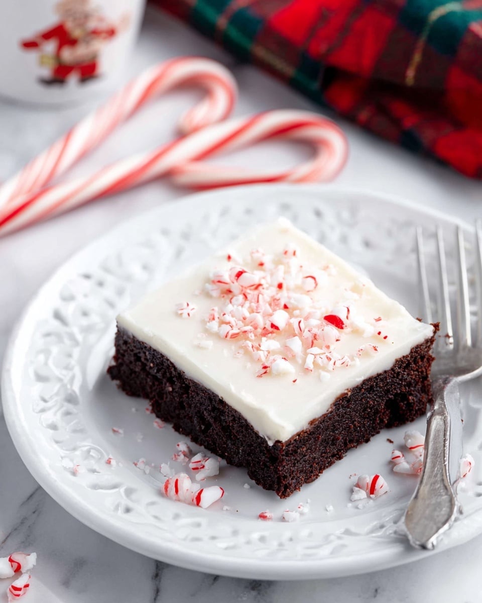 A square piece of dessert sits on a white plate with a delicate floral and pierced design. The dessert has two layers: the bottom is a thick, dark chocolate brownie, and the top is a thick, smooth white frosting sprinkled with small pieces of crushed red and white candy canes. Two candy canes with red and white stripes lay behind the plate on a white marbled surface, along with a few loose candy cane pieces. A silver fork rests on the plate’s edge on the right side. Part of a Santa-themed mug with a red hat is visible to the left. Photo taken with an iphone --ar 4:5 --v 7