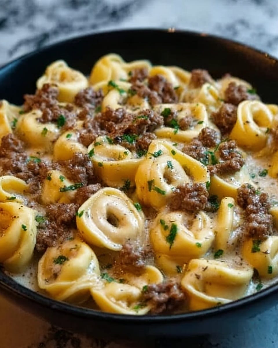 A close-up image of a black bowl filled with tortellini pasta covered in a creamy white sauce with visible small green herb pieces. The tortellini is yellowish with a smooth, slightly shiny texture. On top, there are browned ground meat pieces scattered evenly across the pasta, adding a rich brown color contrast. The surface underneath the bowl is a white marbled texture. The lighting highlights the gloss on the sauce and the edges of the tortellini. photo taken with an iphone --ar 4:5 --v 7