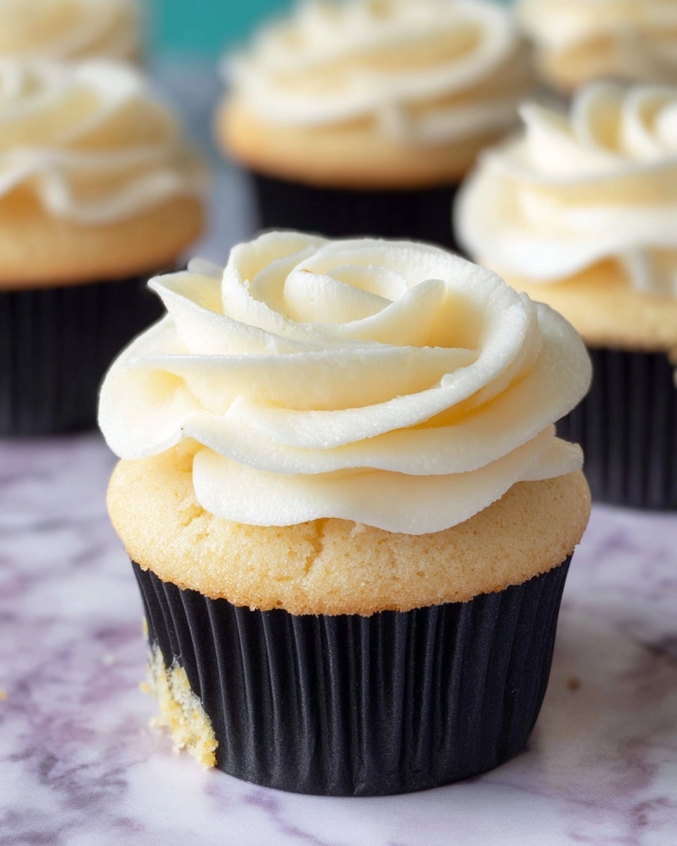 A close-up view of a single vanilla cupcake with a golden-brown base that shows a soft, airy texture. On top is a thick swirl of smooth, white buttercream icing shaped like a rose with visible soft curves and folds. The cupcake is sitting inside a black wrapper with edges flared out slightly, placed on a surface with a white marbled texture and soft pastel colored patterns in the background. Two more similar cupcakes blurred in the background complete the scene. photo taken with an iphone --ar 4:5 --v 7