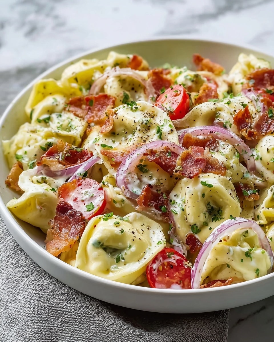 The image shows a white bowl filled with creamy tortellini pasta layered with small pieces of crispy bacon and halved bright red cherry tomatoes. The yellow tortellini have a smooth texture and are coated in a rich white sauce. Thin slices of light red onion and a sprinkle of chopped green herbs are scattered throughout. The dish is finished with a dusting of black pepper and some grated white cheese on top. The bowl sits on a white marbled surface with a soft gray cloth underneath. photo taken with an iphone --ar 4:5 --v 7