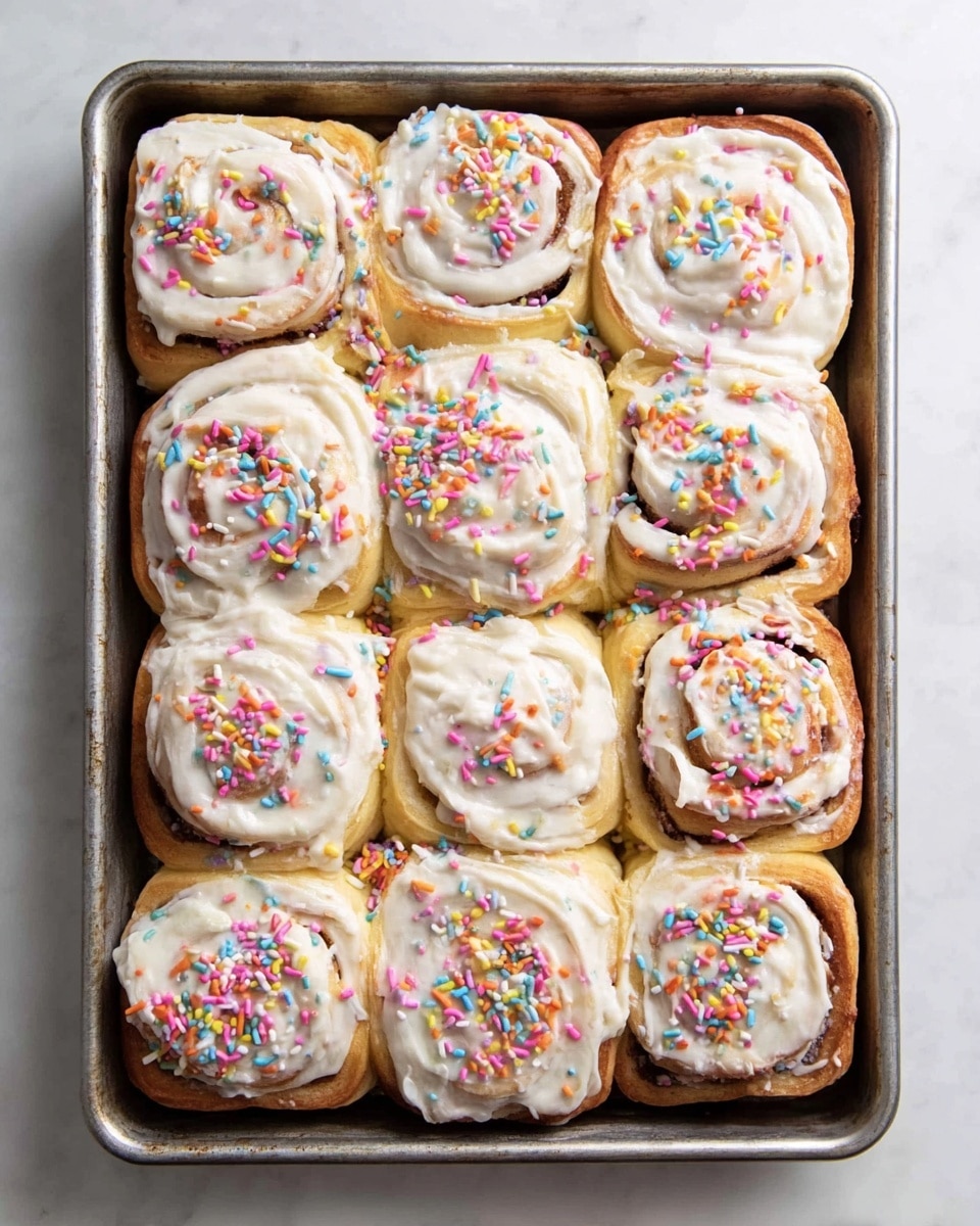 A tray filled with 12 square cinnamon rolls arranged in a 4 by 3 grid, each roll showing soft, golden-brown dough swirled with cinnamon layers and topped thickly with smooth, white cream cheese frosting. Colorful round confetti sprinkles in pink, yellow, green, blue, purple, and orange are scattered evenly over the frosting, adding a bright, festive touch. The tray is set on a white marbled surface, highlighting the warm tones and vibrant colors of the rolls. photo taken with an iphone --ar 4:5 --v 7