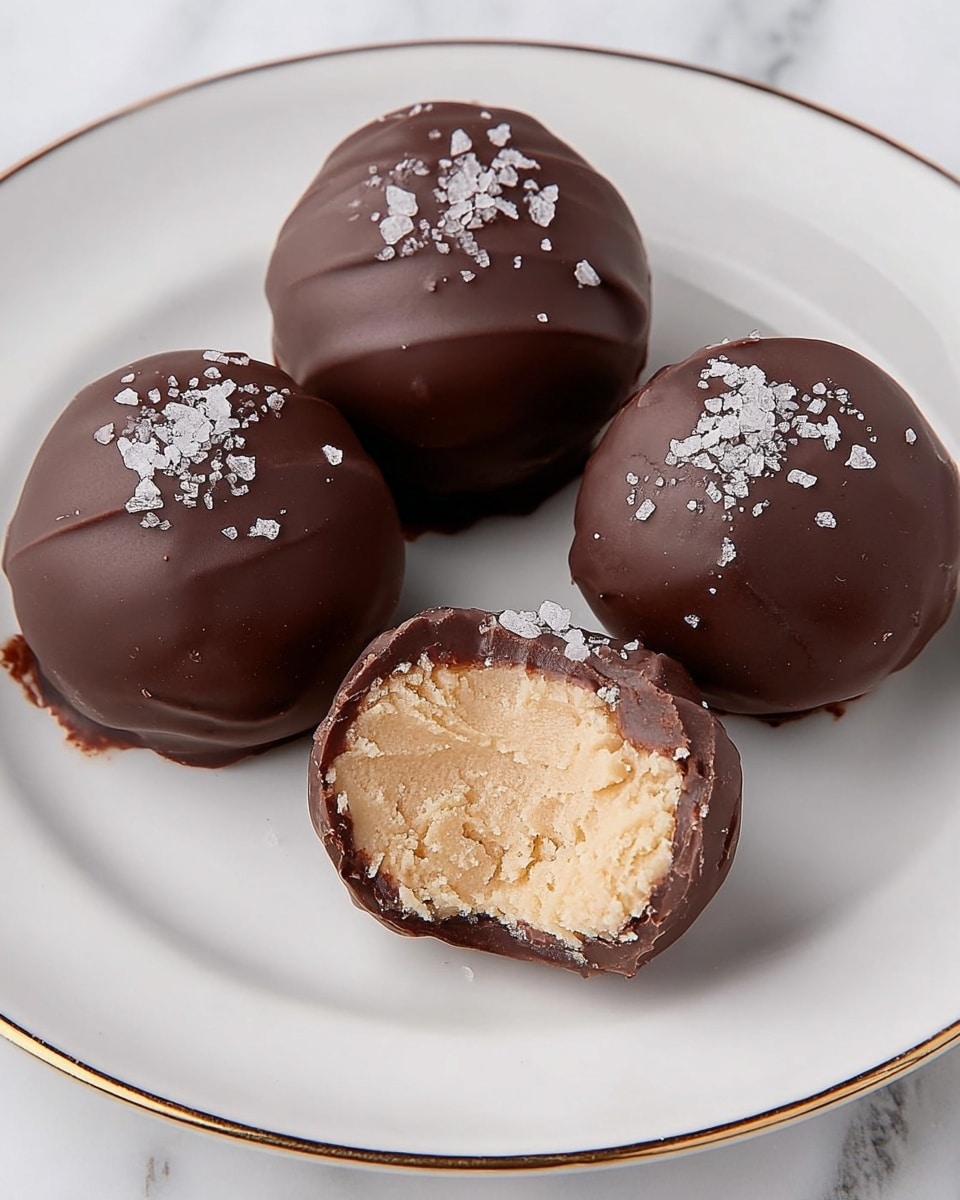 The image shows four round chocolate-covered truffles on a white plate with a thin gold edge, placed on a white marbled surface. Three truffles are whole, with a smooth dark chocolate coating and coarse sea salt crystals sprinkled on top, adding a rough white texture. The fourth truffle is cut in half to display its inside, revealing a creamy, light beige filling with a soft, crumbly texture beneath a thin, glossy layer of dark chocolate shell. The truffles are arranged in a loose cluster, with the cut one in front. Photo taken with an iphone --ar 4:5 --v 7