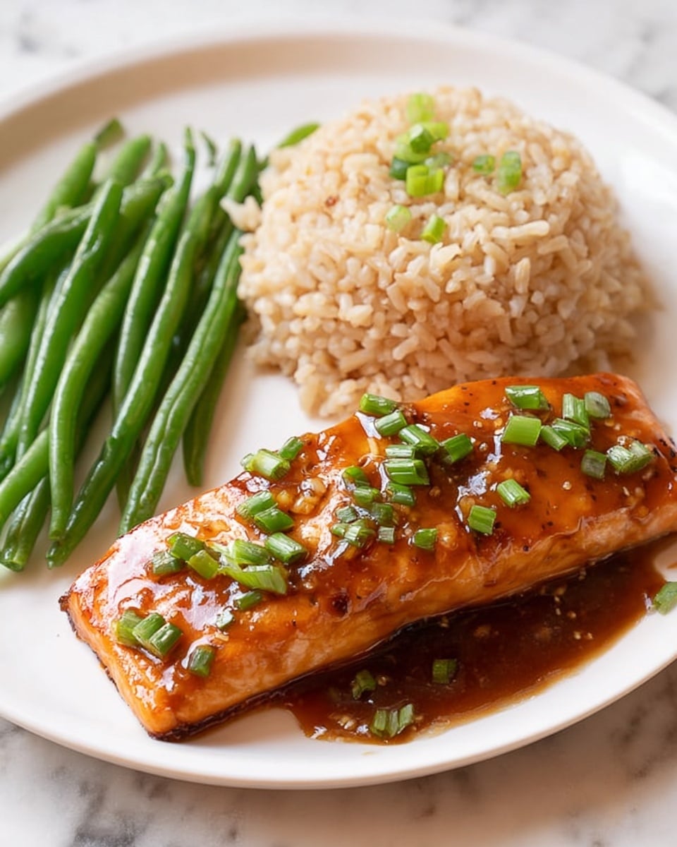 A white plate holds a meal with three main parts. On the right front is a thick, rectangular piece of glazed salmon with a shiny, orange-brown sauce that looks sticky and sweet, topped with small pieces of green onion. Behind the salmon, there is a mound of cooked brown rice, each grain visible and light beige in color, sprinkled with tiny black sesame seeds. On the left side of the plate are several fresh green beans, bright green and smooth. The plate sits on a white marbled surface. photo taken with an iphone --ar 4:5 --v 7