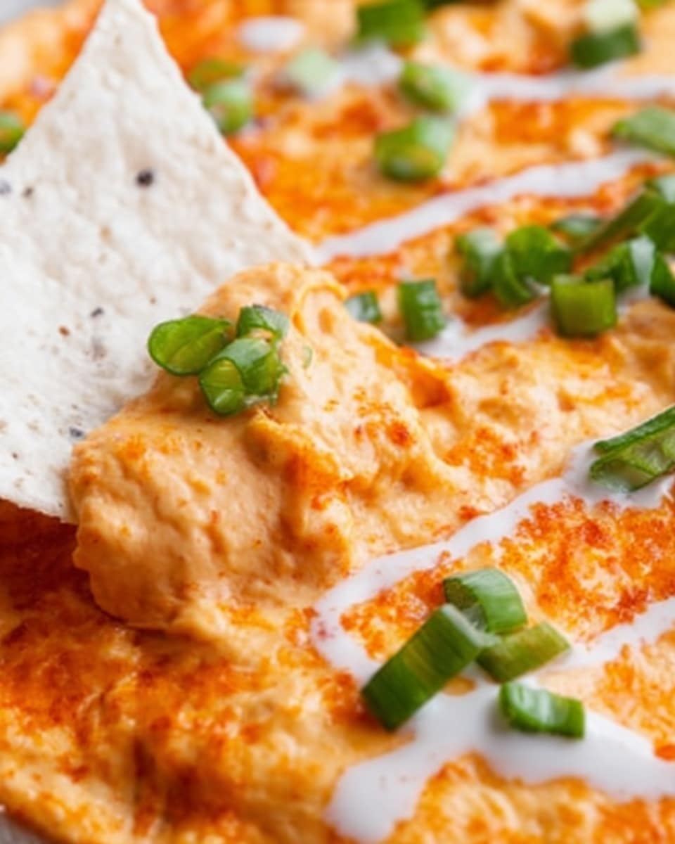 A close-up view of a creamy orange dip topped with small green onion pieces scattered on the surface. A white tortilla chip is partly dipped into the smooth, thick layer of the dip, showing its texture. The background has a white marbled texture, giving a clean and fresh look. photo taken with an iphone --ar 4:5 --v 7