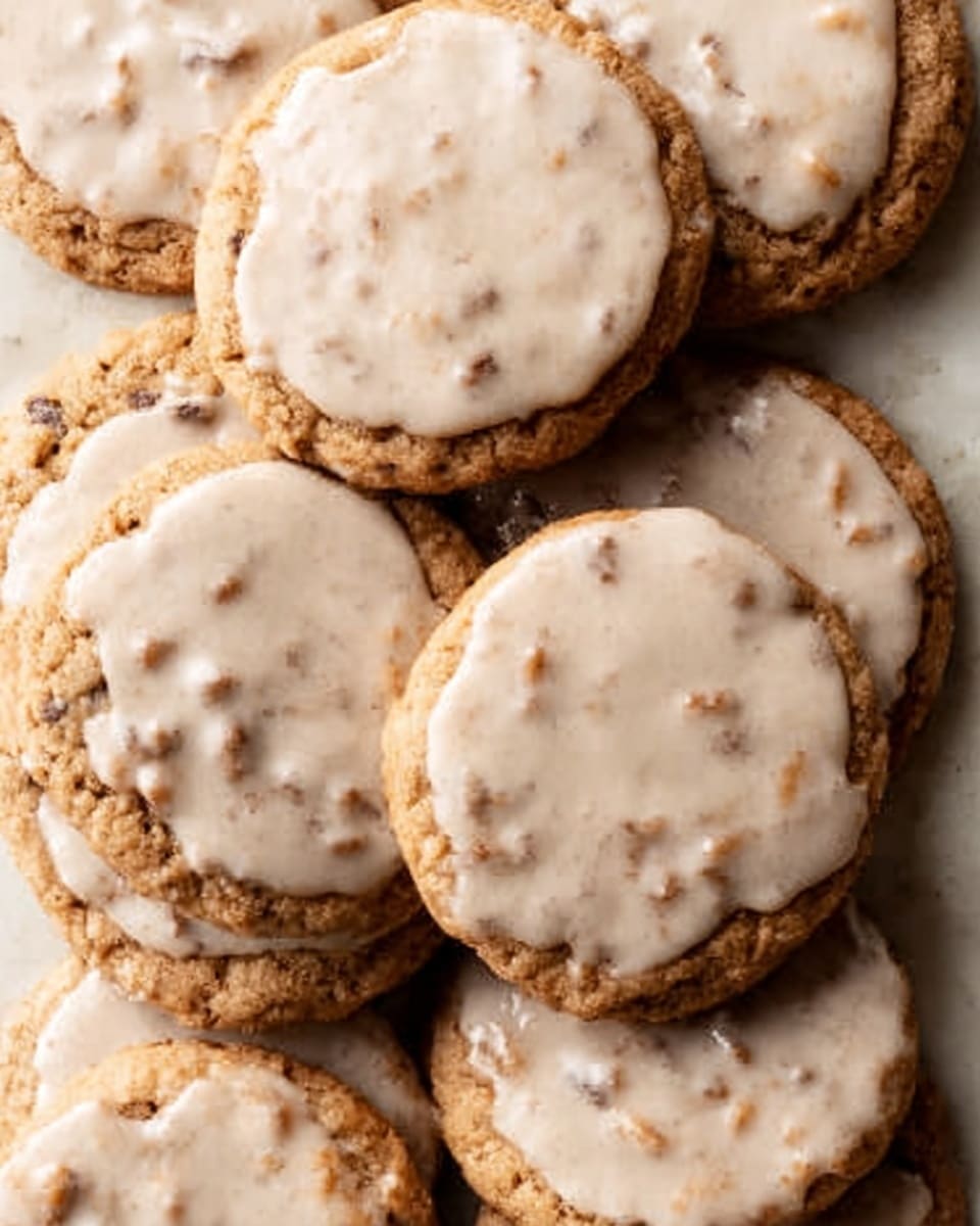 A pile of round cookies with a light brown base color is shown. Each cookie has a smooth, light beige icing layer evenly spread on top, covering nearly the whole surface but leaving slight edges of the cookie visible. The cookies have a slightly rough texture with small chocolate bits embedded within the base, and some variation in size and shape gives them a homemade look. The cookies are arranged closely together on a white marbled surface. photo taken with an iphone --ar 4:5 --v 7
