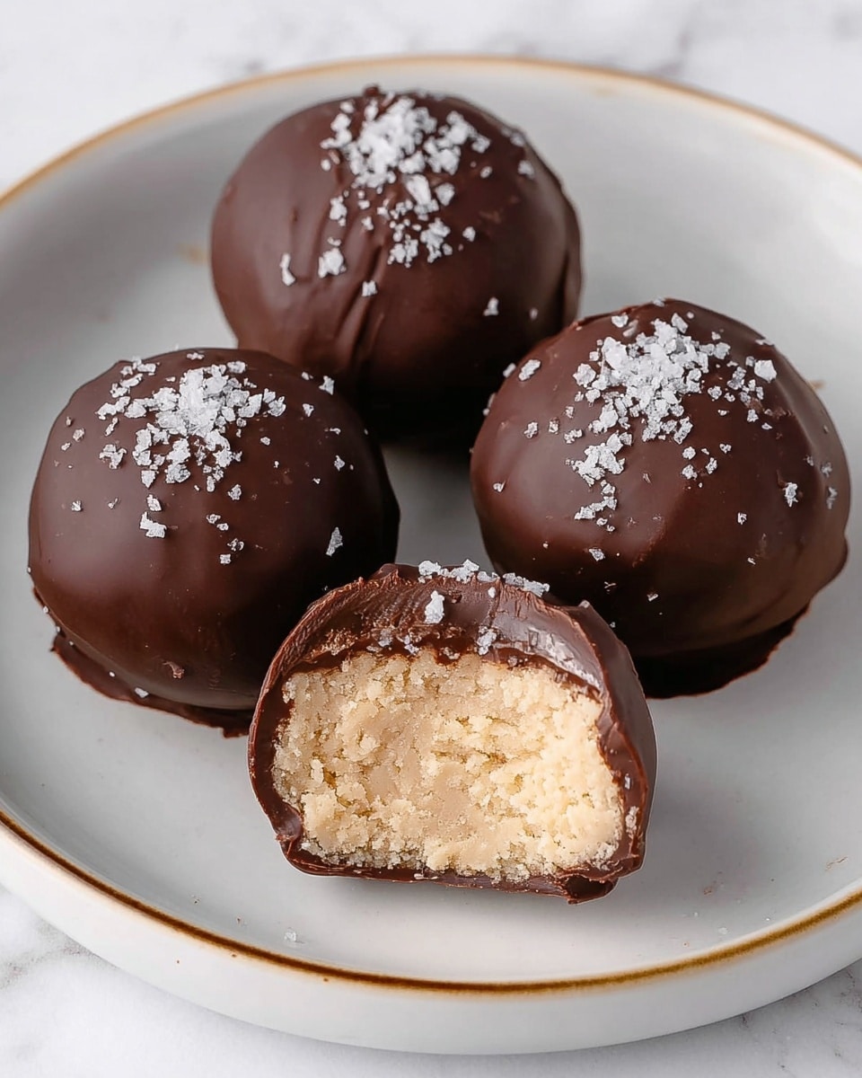 Four round chocolate truffles sit on a white plate with a thin gold rim, placed on a white marbled surface. Three of the truffles are whole, covered in smooth, glossy dark chocolate with small white salt flakes sprinkled on top. The fourth truffle is cut in half, showing a creamy off-white, slightly grainy filling inside surrounded by a thick dark chocolate shell. The truffles are arranged close together toward the center of the plate. photo taken with an iphone --ar 4:5 --v 7