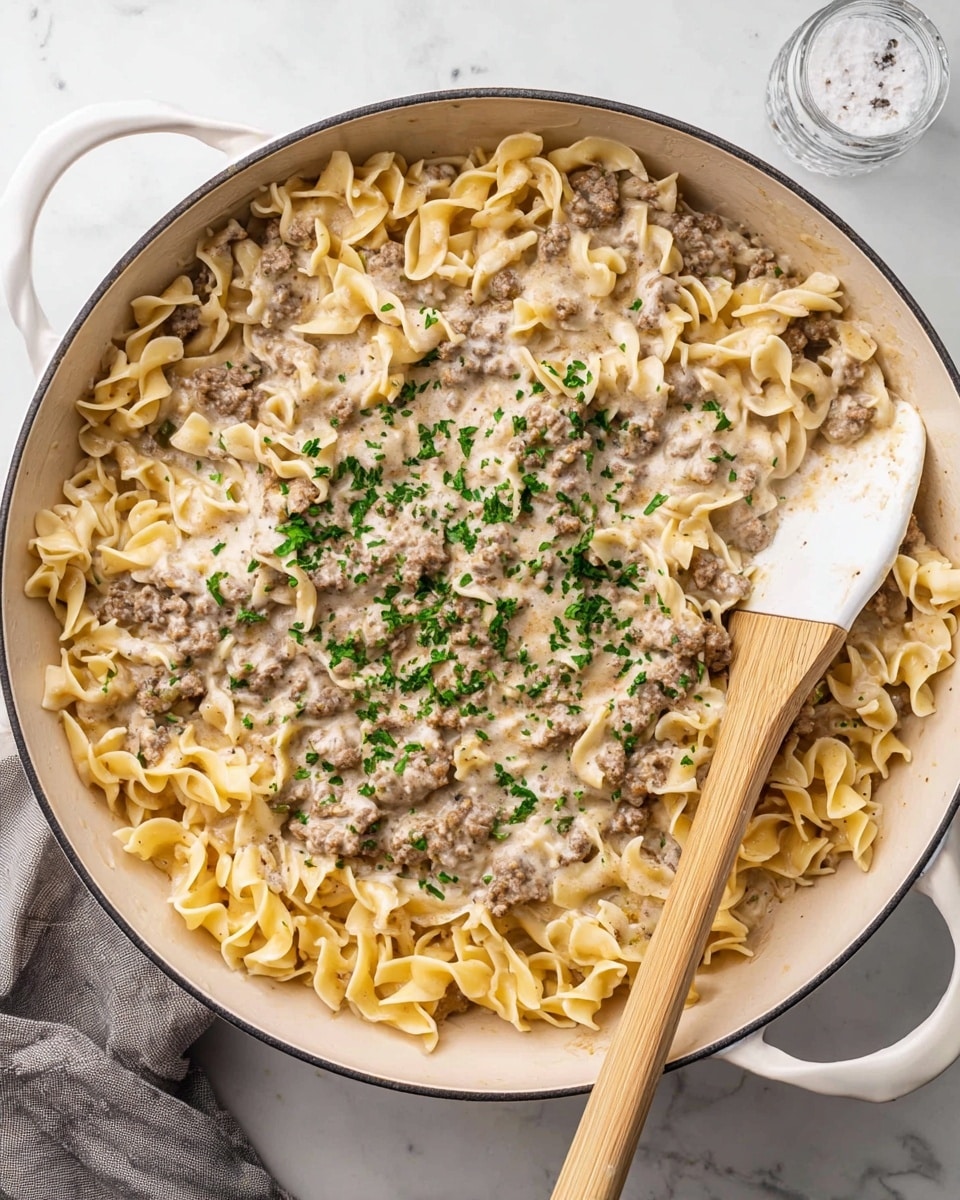A large white skillet filled with a creamy pasta dish made of light yellow egg noodles mixed with small pieces of browned ground beef, all coated in a slightly thick white sauce. Small green parsley pieces are sprinkled evenly across the top, adding color contrast. A white spatula with a light wooden handle is partially inside the skillet on the right side, resting on the noodles. The skillet sits on a white marbled surface, next to a salt shaker and a green patterned cloth. photo taken with an iphone --ar 4:5 --v 7