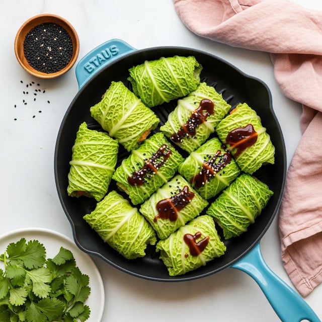 A light blue pan holds twelve green cabbage rolls neatly arranged in a single layer, each roll tightly wrapped with pale green cabbage leaves showing grilled brown marks and white rib veins. The pan rests on a white marbled surface with a small wooden bowl of black seeds at the top. A soft pink cloth is draped softly on the top right corner. Below the pan, on the white marbled surface, there is a white plate with fresh leafy green herbs. Photo taken with an iphone --ar 4:5 --v 7