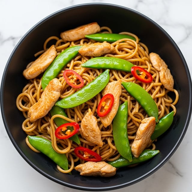 A bowl of stir-fried noodles with pieces of cooked chicken, bright green snap peas, and thin strips of red bell peppers mixed evenly throughout. The noodles are glossy and brown, showing they are coated in a savory sauce. The chicken pieces are light brown and tender-looking, scattered on top and inside the noodles. The snap peas add a fresh green color, while the red peppers provide a bright contrast. The dish is served in a white bowl, sitting on a white marbled surface. Photo taken with an iphone --ar 4:5 --v 7