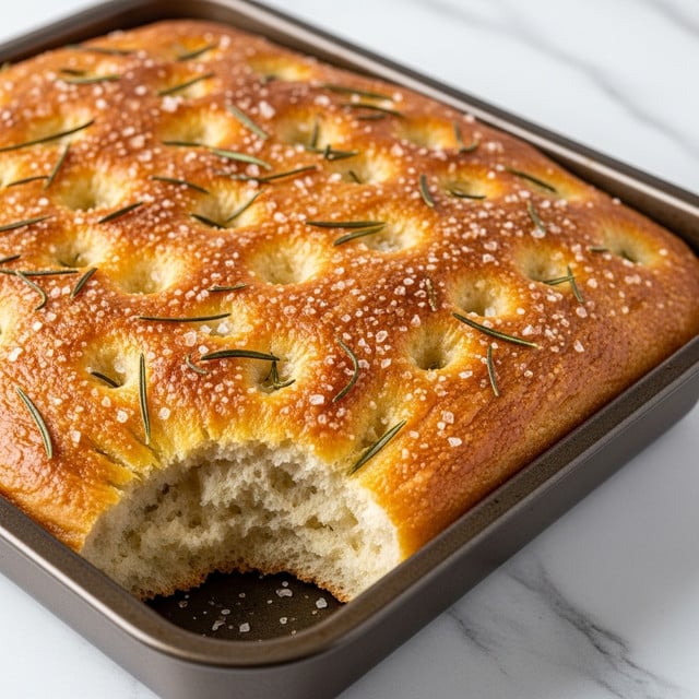 The image shows a golden-brown focaccia bread baked in a dark brown rectangular pan. The top layer is fluffy and shiny, covered with scattered rosemary leaves and a light sprinkling of coarse salt, giving the surface a textured look with small dimples. The bread looks soft and airy inside, visible at the torn edge in the foreground. The background is a white marbled surface, enhancing the warm tones of the bread. Photo taken with an iphone --ar 4:5 --v 7