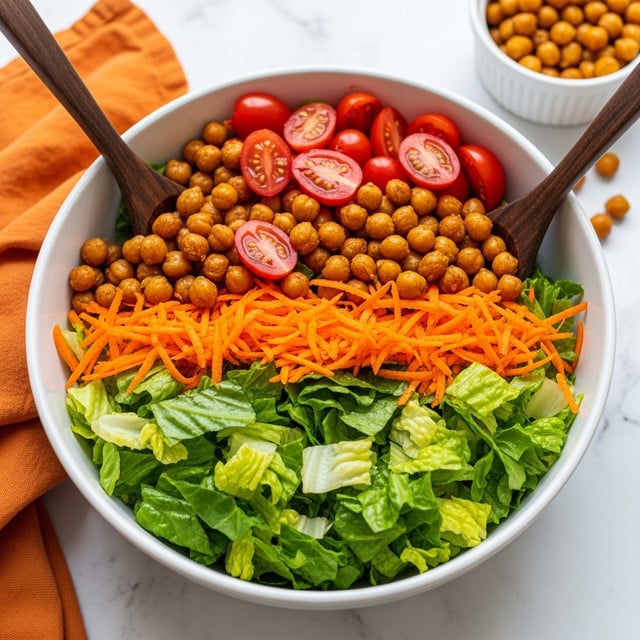A white bowl filled with a mixed salad showing three main layers: the bottom mainly made of shredded green romaine lettuce leaves, the middle layer scattered with orange grated carrots and golden roasted chickpeas, and the top layer dotted with halved roasted cherry tomatoes. Two dark wooden salad spoons rest inside the bowl, partially buried in the salad. The bowl is placed on a white marbled surface with a soft orange cloth nearby, and blurred bowls of roasted chickpeas and other items can be seen in the background. Photo taken with an iphone --ar 4:5 --v 7