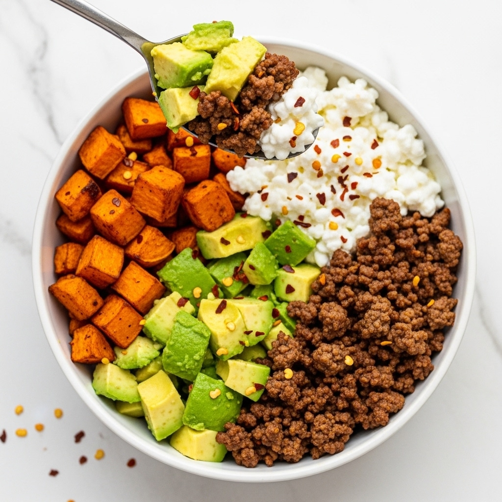A white bowl contains a colorful dish divided into four main sections: bright green avocado chunks on the right, crumbly brown ground beef on the bottom right, golden orange roasted sweet potato cubes on the bottom left, and a white creamy mixture with a slightly lumpy texture sprinkled with red chili flakes on the top left. A silver spoon holds a bite-sized mix of avocado, ground beef, roasted sweet potato, and the creamy white layer above the bowl. The scene is set on a white marbled surface with some scattered red chili flakes. Photo taken with an iphone --ar 4:5 --v 7