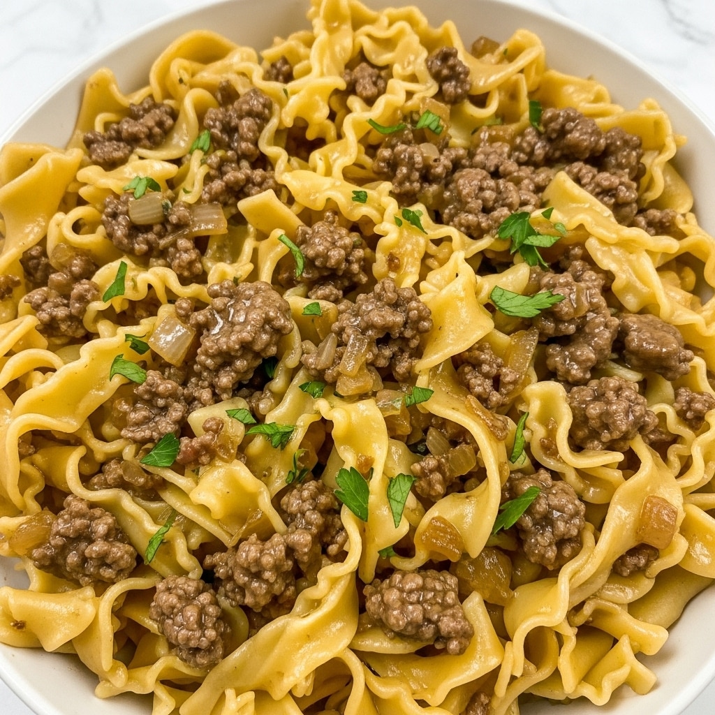This close-up image shows a dish of wide, curly yellow noodles mixed with small chunks of brown cooked ground meat, all coated in a light brown sauce giving a glossy and slightly thick texture. Small green parsley leaves are scattered evenly over the noodles and meat, adding contrast to the warm colors. The noodles have a soft and tender look, with some edges curled and others flat. The sauce pools slightly around the ingredients, with visible black pepper flakes and tiny diced onions throughout the mix. The dish is served in a white bowl placed on a white marbled surface. photo taken with an iphone --ar 4:5 --v 7