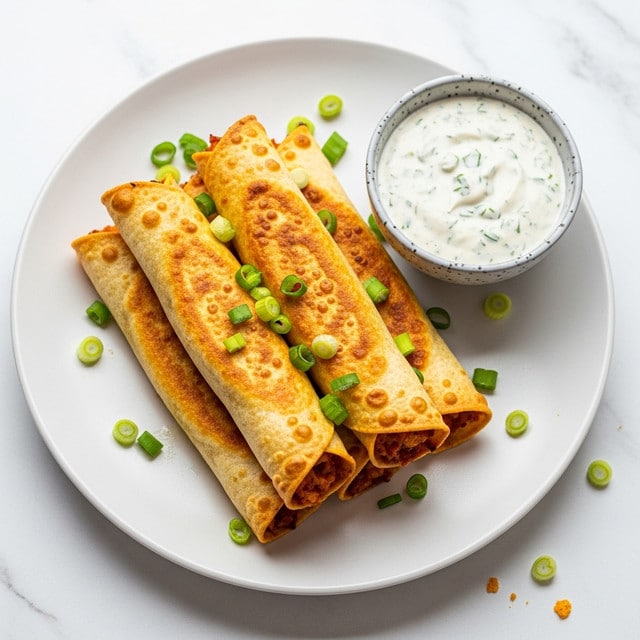 A white plate with five golden brown, crispy rolled taquitos stacked in two layers, showing a lightly bubbled texture on the surface. The taquitos are sprinkled with small, bright green chopped scallions scattered both on top and around them. To the right of the taquitos, a small bowl with a speckled gray rim contains creamy white dipping sauce with green herb flecks. The plate sits on a white marbled surface with a few crumbs and scallion pieces near the edge. Photo taken with an iphone --ar 4:5 --v 7