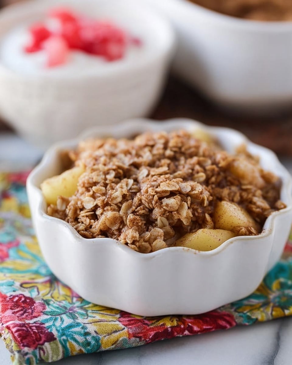 A white scalloped ceramic bowl filled with a three-layered apple crisp dessert: the bottom layer is light golden cooked apple chunks, the middle layer is a soft cinnamon-spiced oat mixture, and the top layer is a crunchy golden brown oat crumble with some visible cinnamon specks. The bowl sits on a colorful floral cloth over a white marbled surface, with a blurred white patterned bowl of yogurt with red fruit pieces in the background. The colors are warm and inviting, with textures ranging from soft fruit to crumbly topping, photo taken with an iphone --ar 4:5 --v 7