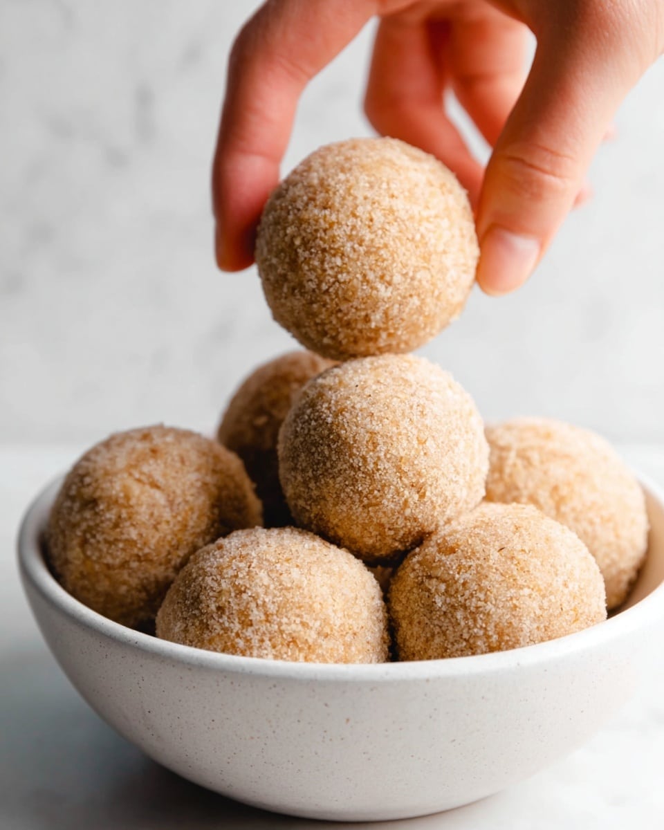 A close-up view of a white bowl filled with round, light brown dough balls that have a rough, grainy texture. A woman's hand is gently holding one of the dough balls above the bowl. The background features a white marbled texture. The image has soft lighting, highlighting the natural details of the dough balls. photo taken with an iphone --ar 4:5 --v 7