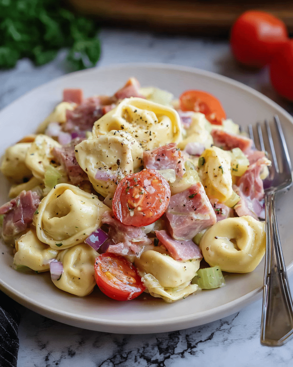 A close-up of a white plate filled with a creamy pasta salad, featuring about three layers of ingredients mixed together: the bottom layer has plump tortellini pasta in a light yellow color with smooth, slightly glossy texture; scattered within and on top are bright red cherry tomato halves with a juicy, shiny surface; pieces of pinkish ham and darker slices of cured meat add a meaty texture; small chunks of pale green celery and bits of finely chopped purple onions add crunch and color contrast; the whole dish is lightly sprinkled with coarse black pepper, enhancing its fresh and hearty look. A silver fork rests on the right side of the plate, placed on a white marbled textured surface with blurred green herbs and a whole cherry tomato in the background. photo taken with an iphone --ar 4:5 --v 7
