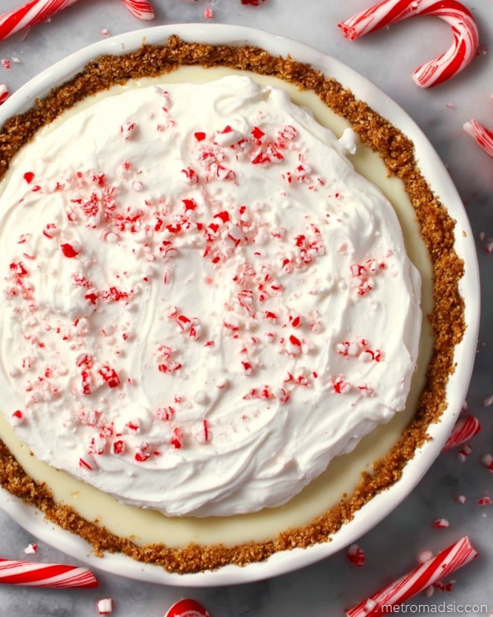 The image shows a pie in a white round dish with a golden brown crust around the edges. The pie has two layers: the bottom layer is a smooth cream filling, and the top layer is a thick, white whipped cream spread evenly with gentle swirls. On top of the whipped cream, there are small red and white crushed peppermint candy pieces sprinkled all over. The dish sits on a white marbled surface with whole peppermint sticks scattered around it. photo taken with an iphone --ar 4:5 --v 7