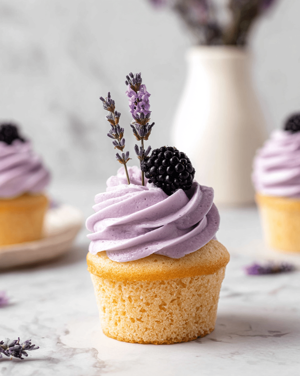 The image shows a single cupcake with a golden yellow base that has a soft, slightly crumbly texture. On top is a thick swirl of light purple frosting with a smooth and creamy texture, stacked in about three to four layers. The frosting is decorated with a small black blackberry and two sprigs of purple lavender flowers standing upright. The cupcake rests on a white marbled surface with a soft-focused background showing another cupcake and a white vase. Photo taken with an iphone --ar 4:5 --v 7