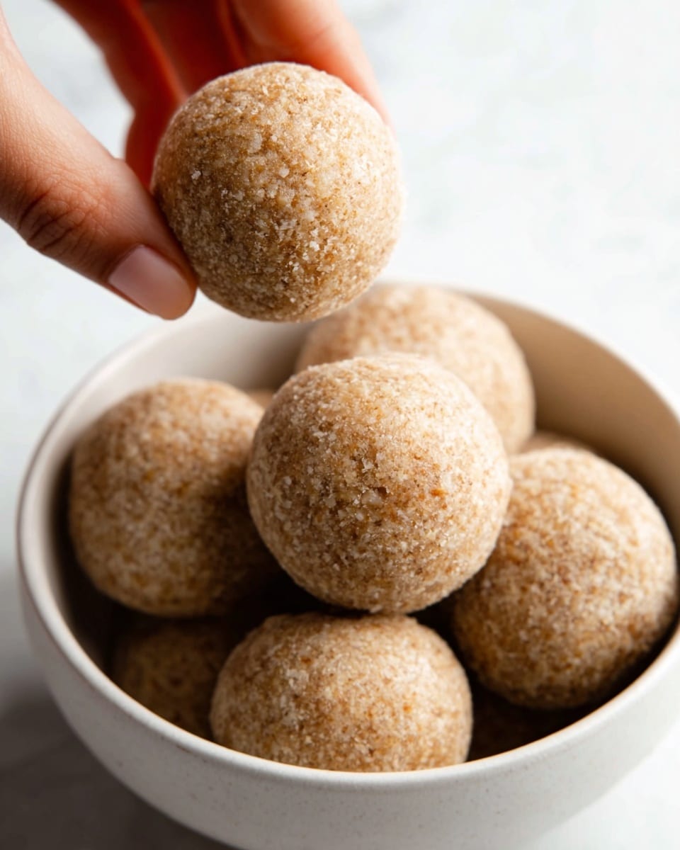 The image shows a close-up of seven round balls with a rough, crumbly texture that looks like they are made from oats or whole grains. They are light brown in color and arranged stacked in a white bowl, with one ball being held above the others by a woman's hand. The background is a white marbled texture, giving a clean and bright look. The surface and lighting highlight the textured surfaces and natural color of the balls clearly. photo taken with an iphone --ar 4:5 --v 7