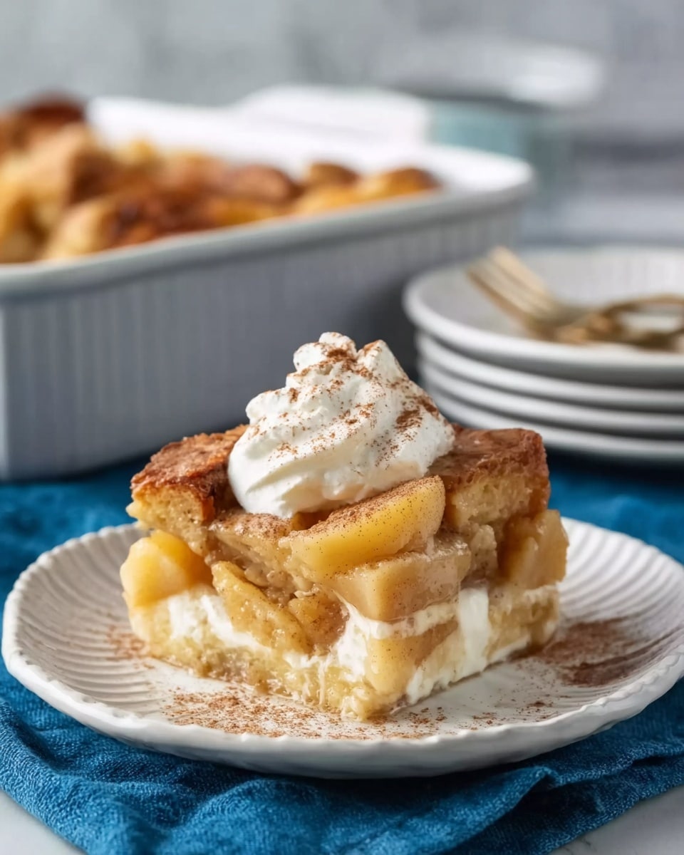 A single serving of bread pudding sits on a white plate with light ridges along the edge, placed on a blue cloth over a white marbled surface. The bread pudding has multiple layers of soft, golden-brown toasted bread soaked in a creamy custard, visible with chunks of apple inside. On top, there is a dollop of whipped cream sprinkled with a light dusting of cinnamon, adding a touch of texture and warmth to the soft dessert. In the background, a white baking dish with more bread pudding and blurred plates and forks can be seen. Photo taken with an iphone --ar 4:5 --v 7