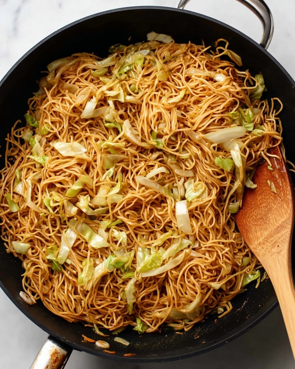 A large black pan filled with loosely piled cooked noodles that are light brown and shiny, mixed evenly with pale green and white chopped vegetable pieces, mostly thin cabbage and sliced green onions; a wooden spatula rests on the upper right side of the pan, slightly touching the noodles, and everything sits on a white marbled surface. photo taken with an iphone --ar 4:5 --v 7