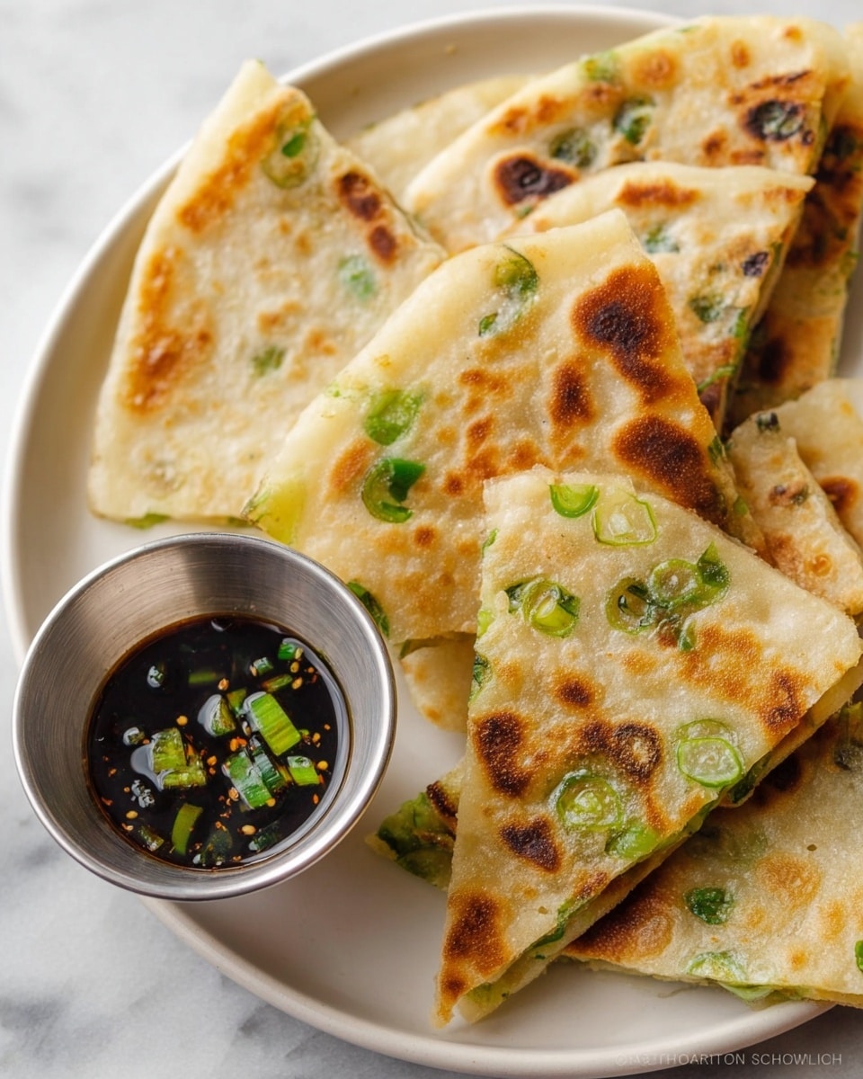 The image shows a close-up of several folded, golden-brown scallion pancakes with visible green onion pieces inside and on top, arranged on a white plate. The pancakes have a slightly crispy texture with scattered dark brown spots from cooking, and some green onion rings are also placed around them. On the right side of the plate, there is a small metal cup filled with dark dipping sauce containing chopped green onions, all set on a white marbled surface. photo taken with an iphone --ar 4:5 --v 7
