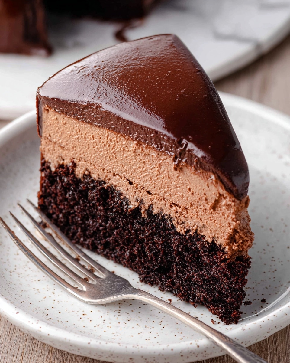 A slice of rich chocolate cake with two layers is shown on a white speckled plate with a silver fork beside it. The bottom layer is a dark, moist chocolate cake with a rough texture, while the top layer is a smooth, creamy chocolate mousse with a lighter brown color. The slice is covered in a glossy, thick chocolate ganache that smoothly coats the side and top edges, creating a shiny surface. The background is a white marbled texture. Photo taken with an iphone --ar 4:5 --v 7