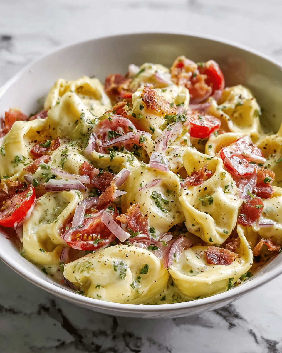 A white bowl filled with creamy tortellini pasta, where each piece is plump and pale yellow with a smooth, soft texture. Mixed in are bright red cherry tomato halves and crispy brown bacon bits scattered throughout. Thin slices of light purple onion add contrast, while finely chopped green herbs are sprinkled on top. The creamy sauce coats the pasta evenly, with specks of black pepper visible across the dish. The bowl sits on a white marbled surface. photo taken with an iphone --ar 4:5 --v 7