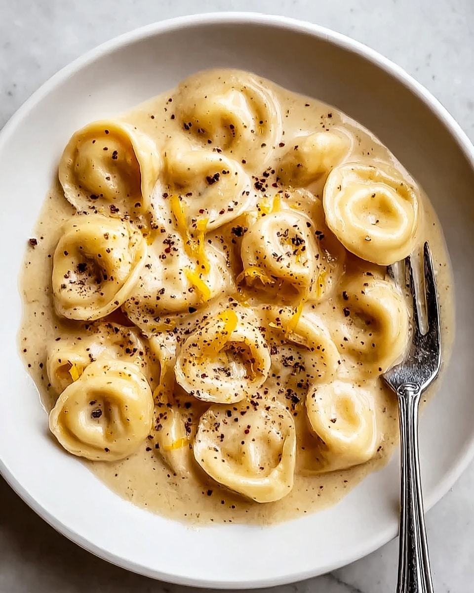A white plate holds a creamy beige sauce with black pepper and small yellow cheese strips mixed evenly. On top are about twelve tortellini pasta pieces, showing their round shape and hollow centers, covered in the smooth sauce. A silver fork rests on the right side of the plate touching the food. The background is a white marbled texture. photo taken with an iphone --ar 4:5 --v 7