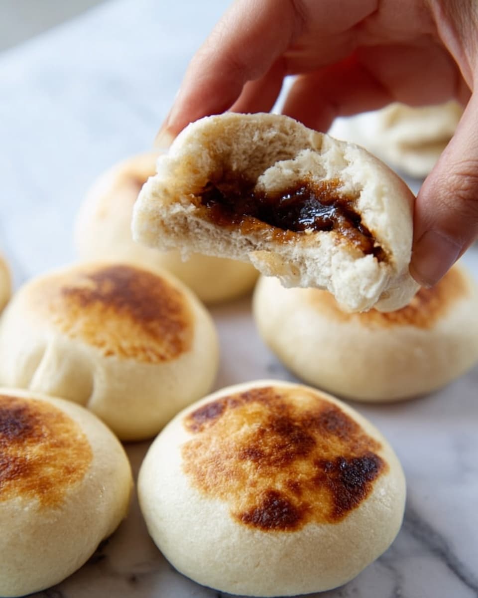 A close-up of a woman's hand breaking apart a small round bun with a soft, light beige outer layer that shows a slightly browned, toasted spot on top. Inside, there is a thick, dark brown filling with a smooth texture. Several other similar buns lie on a white marbled surface in the background, showing their rounded shapes and toasted spots on the top side. Photo taken with an iphone --ar 4:5 --v 7