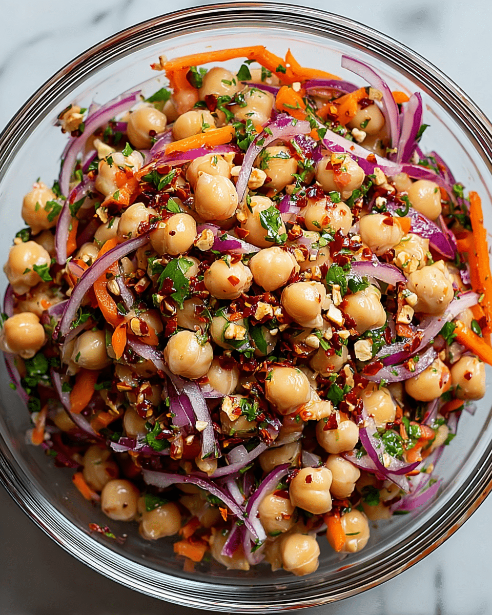A clear glass bowl filled with a colorful salad made of three main layers: a base layer of light beige chickpeas, mixed with thin slices of bright purple-red onion, thin orange carrot strips, and small green chopped herbs scattered evenly throughout. Tiny bits of red chili flakes and crushed nuts or seeds are sprinkled on top, adding texture and contrast. The bowl sits on a smooth white marbled surface, and the salad looks fresh and juicy with a slight shine from dressing. photo taken with an iphone --ar 4:5 --v 7