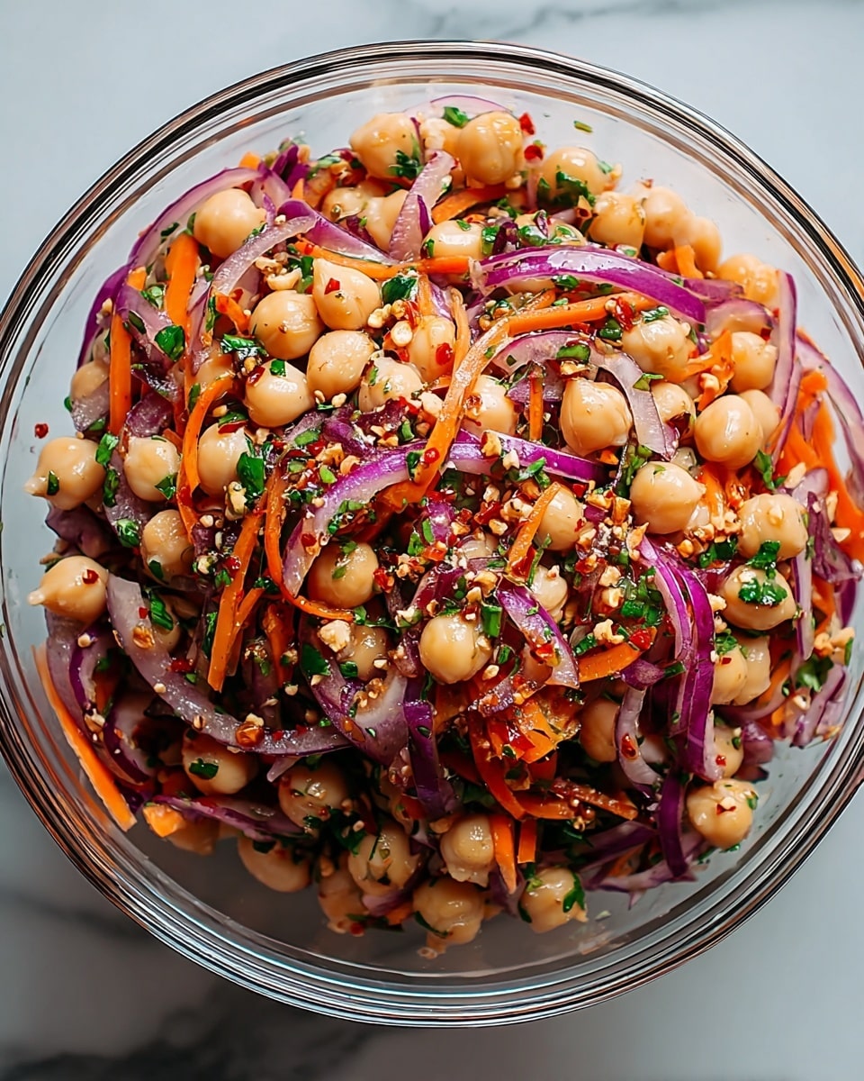 A clear glass bowl filled with a colorful chickpea salad sits on a white marbled texture. The salad has three main layers: at the bottom and through the bowl are creamy beige chickpeas, mixed throughout with thin slices of bright purple-red onions and thin orange carrot strips. Bright green chopped herbs and small bits of crushed red pepper flakes and nuts are scattered evenly, adding texture and color contrast. The salad looks fresh and slightly shiny as if tossed in a light dressing. photo taken with an iphone --ar 4:5 --v 7